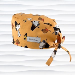 May include: A mustard yellow scrub cap featuring a repeating pattern of Snoopy wearing a pilgrim hat, Woodstock, and roasted turkeys. A rectangular grey fabric label with the text JensCaps is sewn onto the front of the cap.