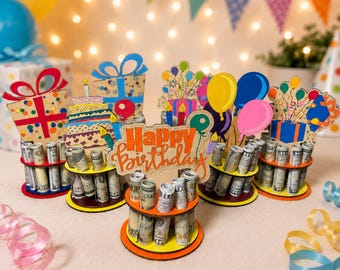 Puede incluir: Un conjunto de cinco soportes de madera para dinero de cumpleaños con recortes coloridos de regalos, pasteles y globos. Cada soporte tiene niveles circulares con orificios para billetes enrollados. La pieza central muestra el texto Happy Birthday.
