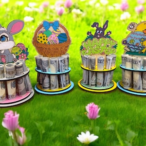 May include: Four Easter-themed money holders with decorative wooden cutouts of a bunny, basket of eggs, a bunny with "Happy Easter" text, and a chick. Each holder is a tiered design with rolled-up bills.