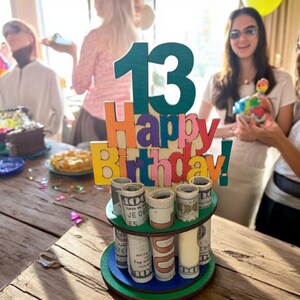 Może przedstawiać: Drewniana dekoracja na tort urodzinowy z numerem 13 w kolorze morskim i napisem "Happy Birthday!" w kolorowych literach. Ciasto ma dwa piętra z rolowanymi banknotami. Balony są w tle.
