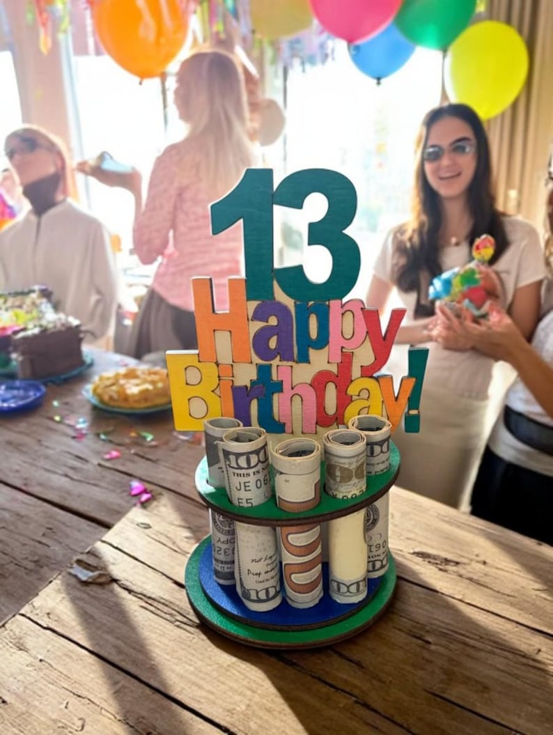 Może przedstawiać: Drewniana dekoracja na tort urodzinowy z numerem 13 w kolorze morskim i napisem "Happy Birthday!" w kolorowych literach. Ciasto ma dwa piętra z rolowanymi banknotami. Balony są w tle.