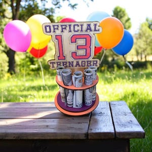 Może przedstawiać: Drewniany uchwyt na prezent urodzinowy z napisem "OFFICIAL 13 TEENAGER" w kolorze fioletowym i czerwonym. Uchwyt ma trzy poziomy zrolowanych banknot&oacute;w. Kolorowe balony w tle.