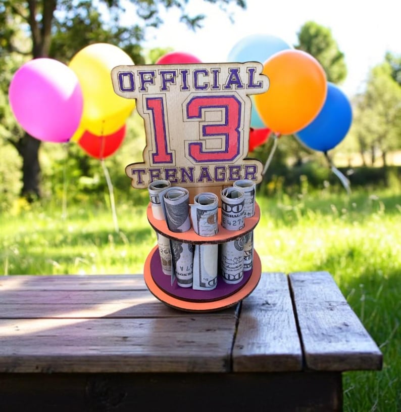 Może przedstawiać: Drewniany uchwyt na prezent urodzinowy z napisem "OFFICIAL 13 TEENAGER" w kolorze fioletowym i czerwonym. Uchwyt ma trzy poziomy zrolowanych banknot&oacute;w. Kolorowe balony w tle.