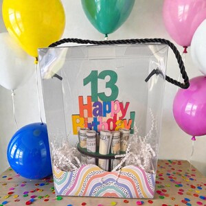 Może przedstawiać: Przezroczyste pudełko na prezent z czarnym uchwytem ze sznurka, wypełnione zwiniętymi banknotami i białym papierem. Pudełko ma kolorowy znak "Happy Birthday!" z numerem "13". Kolorowe balony i konfetti otaczają pudełko.