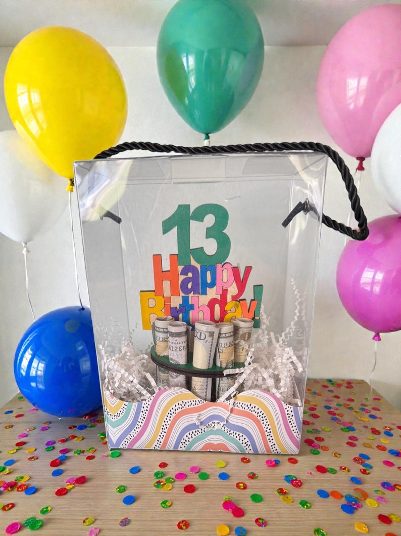 Może przedstawiać: Przezroczyste pudełko na prezent z czarnym uchwytem ze sznurka, wypełnione zwiniętymi banknotami i białym papierem. Pudełko ma kolorowy znak "Happy Birthday!" z numerem "13". Kolorowe balony i konfetti otaczają pudełko.