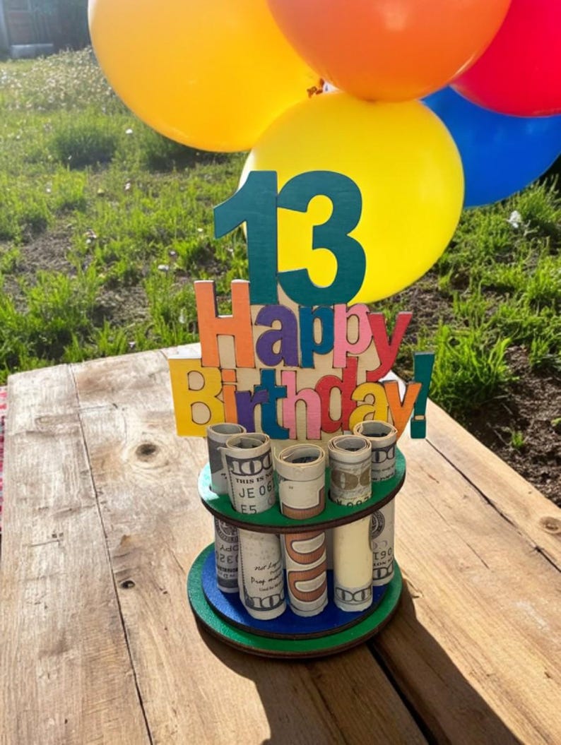 Może przedstawiać: Tort urodzinowy z kolorowym napisem "Happy Birthday!" i liczbą "13". Tort jest ozdobiony zwiniętymi banknotami dolara. Balony w kolorze ż&oacute;łtym, pomarańczowym i niebieskim w tle. Tort stoi na drewnianym stole.