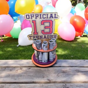 Może przedstawiać: Drewniany uchwyt na pieniądze z napisem "OFFICIAL 13 TEENAGER" w kolorze czerwonym i fioletowym. Uchwyt ma trzy poziomy zrolowanych banknot&oacute;w. Kolorowe balony w tle, a uchwyt stoi na drewnianej powierzchni.