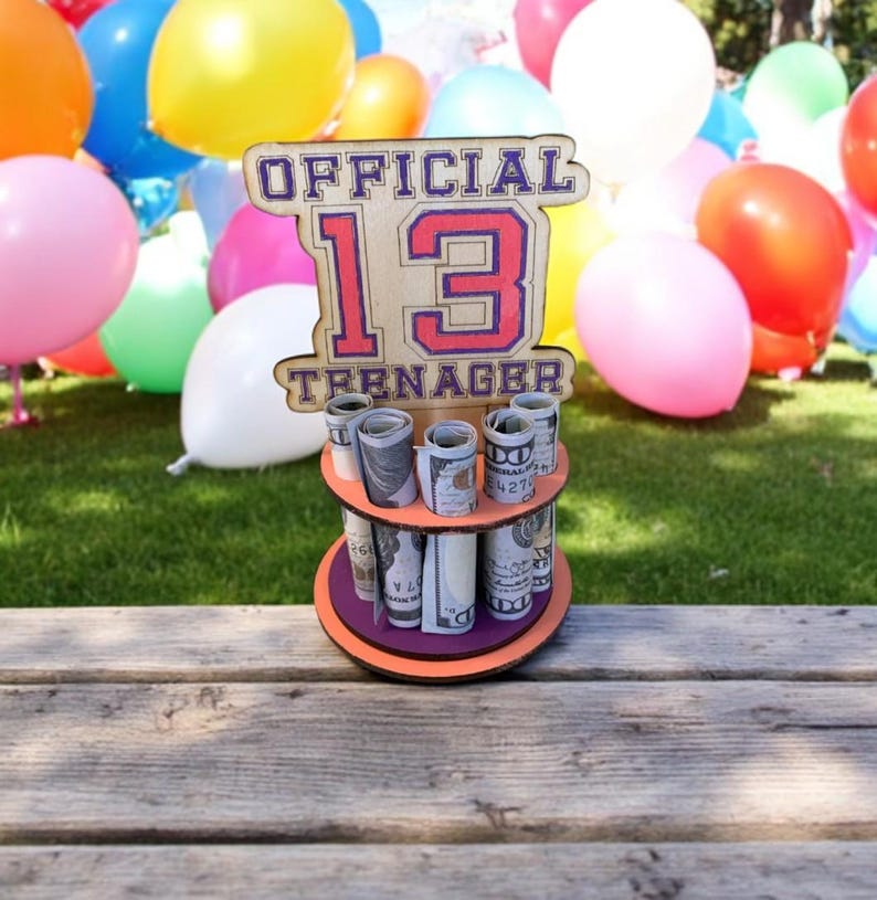 Może przedstawiać: Drewniany uchwyt na pieniądze z napisem "OFFICIAL 13 TEENAGER" w kolorze czerwonym i fioletowym. Uchwyt ma trzy poziomy zrolowanych banknot&oacute;w. Kolorowe balony w tle, a uchwyt stoi na drewnianej powierzchni.
