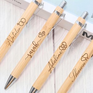 May include: Four bamboo pens with silver tips, each engraved with a name and a small design. The names "Natalia", "Sarah", "Adeline", and "Shawn" are visible. The pens are arranged on a light-coloured surface.