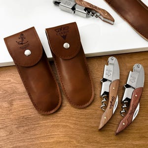 May include: A collection of wine openers with wooden handles and stainless steel components. Some openers are in brown leather cases. The openers have the names "Dalton", "Weglor", and "Carter" engraved on them.