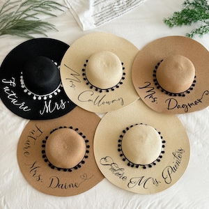 Peut inclure: Cinq chapeaux de paille à larges bords avec des inscriptions noires et des bordures à pompons. Les chapeaux sont noirs, beiges et bruns, avec le texte "Future Mrs." et des noms. Les chapeaux sont disposés sur une surface blanche.