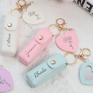 Può includere: Una collezione di portachiavi personalizzati in vari colori, tra cui bianco, rosa e blu. Ogni portachiavi presenta un piccolo astuccio con un nome stampato e un ciondolo a forma di cuore con un disegno floreale o uno specchio. I portachiavi hanno elementi dorati.