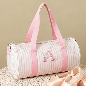 May include: A pink and white striped duffel bag with pink handles and trim, featuring an embroidered letter "A" and a bunny. A pair of pink ballet shoes with ribbons are next to the bag. The bag is on a lace-covered table.