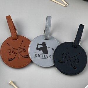 May include: Three personalized golf bag tags in brown, gray, and black. Each tag is round with a strap and features a golf-themed design and a name or initials. Golf tees are scattered around the tags.