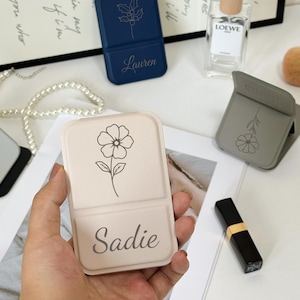 Może przedstawiać: Kolekcja spersonalizowanych stojaków na telefony w różnych kolorach. Jeden jest trzymany w dłoni, z kwiatowym wzorem i imieniem "Sadie". Inne stojaki są granatowe i szare, z kwiatowymi wzorami i imionami "Lauren" i "Tote This."