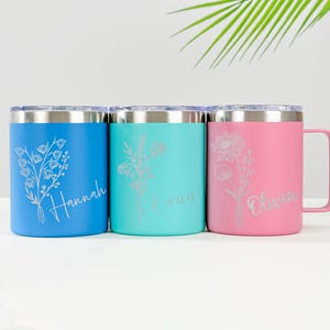 Könnte beinhalten: Drei isolierte Reisebecher in Blau, Türkis und Rosa. Jeder Becher hat ein Blumendesign und einen Namen in Schreibschrift: Hannah, Lauren und Olivia. Die Becher haben silberne Ränder und Griffe.