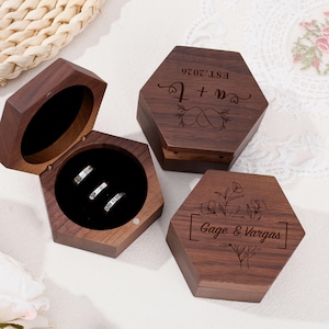 Pode incluir: Três caixas hexagonais de madeira para anéis com detalhes gravados. Uma caixa está aberta, revelando três anéis no interior. As outras duas caixas estão fechadas, com texto gravado e desenhos florais.