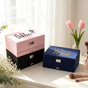 Könnte beinhalten: Drei Schmuckkästchen aus Samt in Rosa, Schwarz und Marineblau. Die rosa Box ist mit "Dahlia" und einem Diamanten-Motiv beschriftet. Die marineblaue Box ist mit "Winnie 12.05.2026" beschriftet. Jede Box hat eine Schublade und einen silbernen Verschluss.