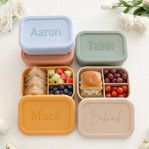 May include: Four rectangular lunch boxes in pastel colors, each with a name embossed on the lid. Two boxes are open, revealing sandwiches, fruit, and other snacks. The boxes are arranged on a white surface, with flowers in the background.