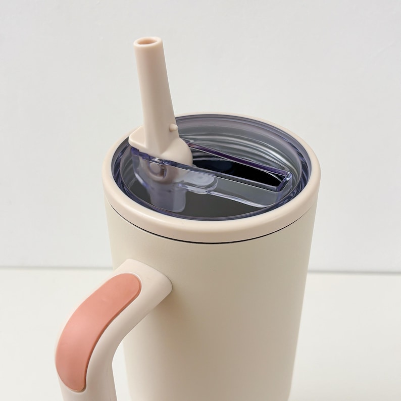 May include: A cream-colored insulated tumbler with a handle and a clear lid. The handle has a peach-colored accent. A matching cream-colored straw extends from the lid. The tumbler is designed to keep beverages cold or hot.