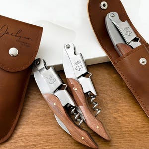 May include: Three wine bottle openers with wooden handles and stainless steel components. The openers are displayed with their brown leather cases. One case is embossed with the name "Jackson" and the text "Groomsman 25.5.2026".