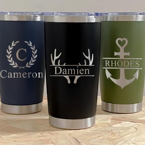Peut inclure: Trois gobelets en acier inoxydable avec couvercles transparents. L'un est bleu marine avec une couronne de laurier et le nom "Cameron". Le second est noir avec un motif de bois de cerf et le nom "Damien". Le troisième est vert olive avec une ancre et le nom "Rhodes".