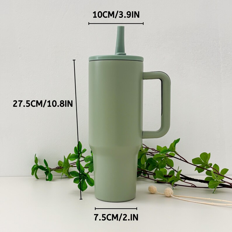 May include: A sage green insulated tumbler with a handle, lid, and straw. The tumbler is 10.8 inches tall, 3.9 inches wide, and 2 inches in diameter. The tumbler is designed to keep beverages cold or hot. Green foliage is in the background.