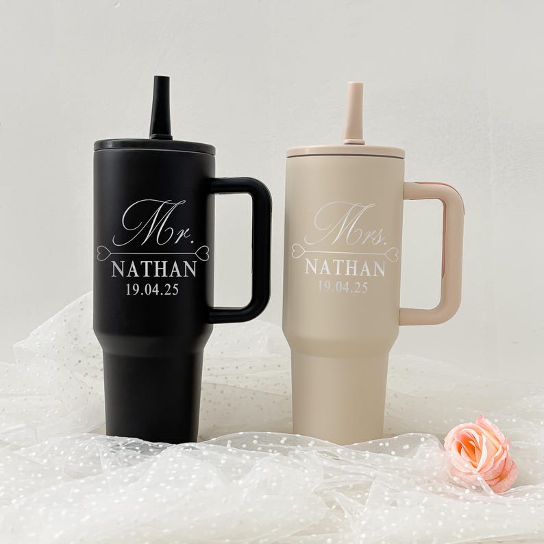 May include: Two insulated tumblers, one black and one beige, with handles and straws. The black tumbler is inscribed with "Mr. NATHAN 19.04.25" in white script. The beige tumbler is inscribed with "Mrs. NATHAN 19.04.25".