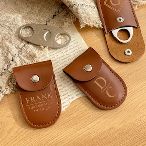 K&ouml;nnte beinhalten: Ein silberner Zigarrenschneider und drei braune Zigarrenetuis aus Leder. Ein Etui ist mit dem Namen "FRANK" und dem Text "GROOMSMEN 04.19.25" gepr&auml;gt. Die anderen Etuis haben Initialen und ein dekoratives Design.