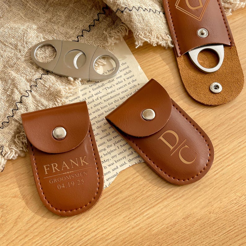 K&ouml;nnte beinhalten: Ein silberner Zigarrenschneider und drei braune Zigarrenetuis aus Leder. Ein Etui ist mit dem Namen "FRANK" und dem Text "GROOMSMEN 04.19.25" gepr&auml;gt. Die anderen Etuis haben Initialen und ein dekoratives Design.