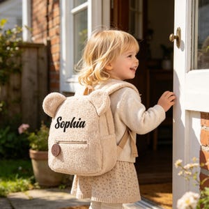 May include: A beige teddy bear backpack with the name "Sophia" printed on the front. The backpack has bear ears, a front zipper pocket, and adjustable straps. The child is wearing a matching sweater and skirt.