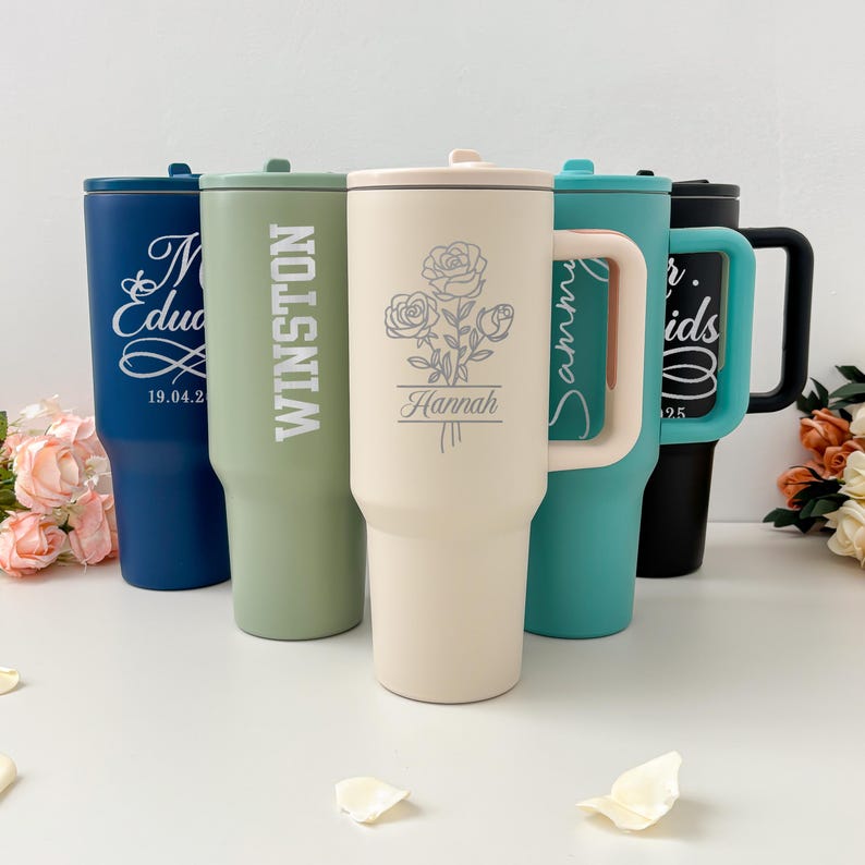 May include: A collection of personalized tumblers in various colors, including blue, green, cream, teal, and black. Each tumbler features custom text and designs, such as names, dates, and floral illustrations. The tumblers have handles and lids.