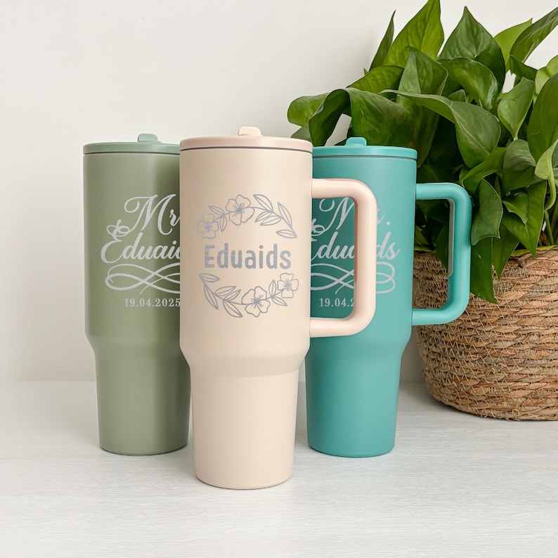 May include: Three insulated tumblers in pastel colors: sage green, pale pink, and teal. Each tumbler features a lid and a handle on the teal one. The pink tumbler has a floral wreath design with the name "Eduaids". The sage green and teal tumblers have the names "Mr Eduaids" and "N Eduaids" with a date.