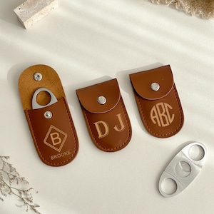 K&ouml;nnte beinhalten: Eine Sammlung brauner Leder-Zigarrenetuis mit personalisierten Initialen und einem silbernen Zigarrenschneider. Ein Etui ist ge&ouml;ffnet und zeigt den Schneider. Die Etuis sind mit den Buchstaben "B", "DJ" und "ABC" gepr&auml;gt. Das Wort "BROOKE" ist ebenfalls sichtbar.