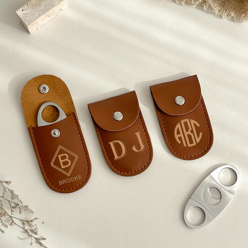 K&ouml;nnte beinhalten: Eine Sammlung brauner Leder-Zigarrenetuis mit personalisierten Initialen und einem silbernen Zigarrenschneider. Ein Etui ist ge&ouml;ffnet und zeigt den Schneider. Die Etuis sind mit den Buchstaben "B", "DJ" und "ABC" gepr&auml;gt. Das Wort "BROOKE" ist ebenfalls sichtbar.