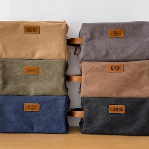 May include: A selection of canvas wash bags in shades of tan, grey, olive green, and navy blue. Each bag has a leather handle and a rectangular leather patch with initials or names.