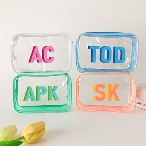 May include: Four clear, rectangular cosmetic bags with colorful initial letters: AC in pink, TOD in blue, APK in green, and SK in yellow and orange. Each bag has a colored border and a zipper closure. The bags are stacked and ready for travel.