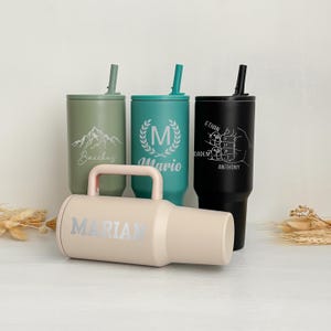 May include: Four personalized tumblers with straws and lids in various colors: olive green, teal, and black. One tumbler is light beige with a handle. Each tumbler has custom text or a graphic, including names and a mountain scene.