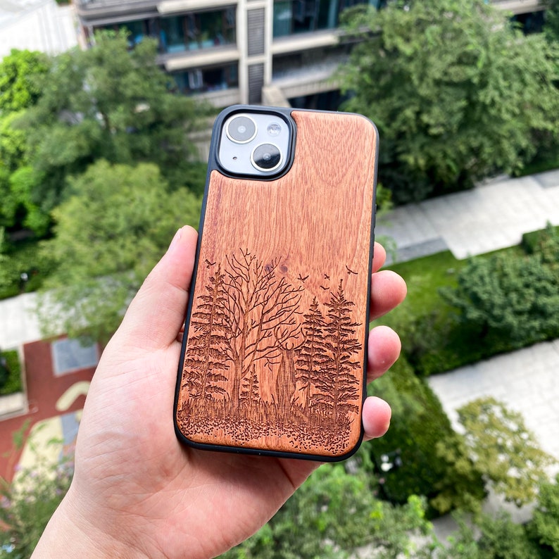 May include: A wooden phone case with a laser-engraved forest scene. The case is brown with a black border and features a design of trees and birds.