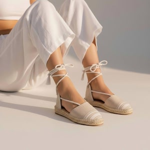 Op de afbeelding: Espadrille sandalen met een beige canvas bovenwerk en een gestreept ontwerp op de neus. De sandalen hebben een geweven jute zool en witte enkelbanden met kwastjes. Schoenen op een wit oppervlak.