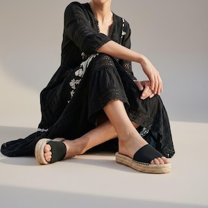 May include: Black espadrille sandals with a thick woven sole and a solid black strap. The sandals are worn with a black dress with white embroidery and lace trim. The dress has long sleeves and a v-neck.