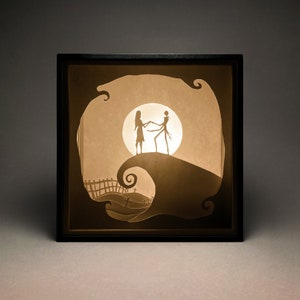 Op de afbeelding: Een zwarte omlijsting met een schaduwdoos met een papier-uitgesneden ontwerp van een stel dat op een heuvel danst. Het stel is gesilhouetteerd tegen een volle maan. De heuvel is een gestileerde weergave van een heuvel met een kronkelend pad.