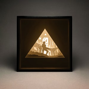 May include: A black framed shadow box with a 3D paper cut-out design of a futuristic scene. The scene features a group of figures in a triangular frame, with a building in the background.