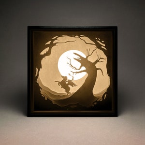 May include: A black framed shadow box with a paper cut-out design of a witch riding a broomstick under a full moon and a large tree. The design is illuminated from behind, creating a glowing effect.