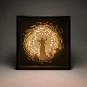 May include: A black shadowbox with a paper cut-out design of a woman standing in a circle of runes. The woman is wearing a long robe and has her arms raised. The background is a dark brown color.