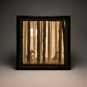 May include: A black framed shadow box with a paper cut-out scene of a wolf and a girl in a red hood walking through a forest. The scene is illuminated from behind, creating a glowing effect.