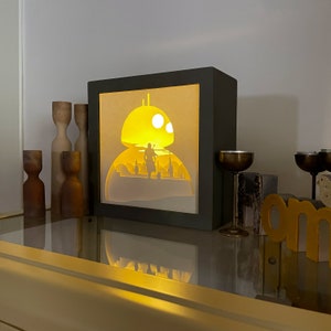May include: A black framed light-up box with a white silhouette of a Star Wars character standing in front of a yellow and white spaceship. The box is lit from behind, creating a glowing effect.