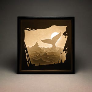 Puede incluir: Una caja de sombra enmarcada en negro con un recorte de papel blanco de una cola de ballena que se eleva de las olas del océano. La cola de ballena está retroiluminada con una luz blanca cálida, creando un efecto brillante.