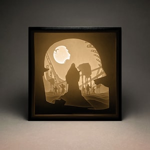 May include: A black framed shadow box with a paper cut-out design of a Star Wars scene. The scene features a hooded figure standing in front of a large moon with a group of figures in the background.