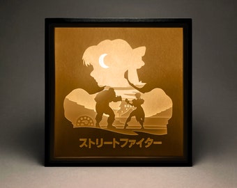 Peut inclure: Une œuvre d'art lumineuse encadrée de noir avec une silhouette de deux personnages se battant. L'arrière-plan est de couleur brun clair avec un croissant de lune. Le texte "Street Fighter" est écrit en japonais en bas de l'image.